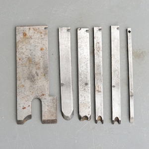 Può includere: Set di sette scalpelli per legno in metallo con diverse forme e dimensioni delle lame. Gli scalpelli sono in metallo argentato con un aspetto leggermente arrugginito.