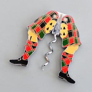 Può includere: Un cavatappi di fantasia a forma di gambe che indossano un kilt scozzese rosso, nero e verde. Il cavatappi è in metallo e ha una finitura argentata. Il testo "PAT. PEND." è visibile sul cavatappi.