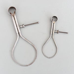 Puede incluir: Dos calibradores antiguos de color plateado de diferentes tamaños. Cada calibrador tiene un extremo curvado en forma de bucle y un mecanismo de ajuste de tornillo. Los calibradores están diseñados para medir el diámetro de un objeto.