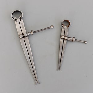 Puede incluir: Dos calibradores antiguos de color plateado, uno grande y otro pequeño, se muestran sobre un fondo gris. Los calibradores tienen una parte superior curvada, brazos ajustables y un mecanismo de tornillo para mediciones precisas. El calibrador más grande tiene texto.