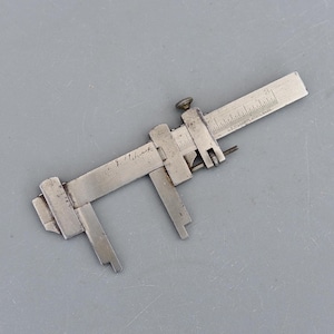 Könnte beinhalten: Ein silberfarbener Vintage-Messschieber mit einer verschiebbaren Skala und einer Schraube zur Einstellung. Das Werkzeug hat geätzte Markierungen und den Text "J. Schrock" und "L.S. Starrett Co. Athol Mass". Der Messschieber ist für präzise Messungen konzipiert.