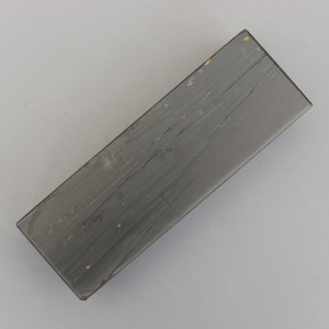 Puede incluir: Una piedra de afilar rectangular gris con arañazos visibles y pequeñas motas doradas. La piedra está ligeramente inclinada, mostrando su forma rectangular y superficie desgastada. Esta herramienta se utiliza para afilar cuchillos y otras hojas.