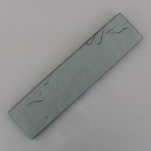 May include: A rectangular, grey-green glass tile with a textured surface. The tile has a slightly irregular shape and appears to have some markings or imperfections. The tile is angled, showing its length and width.
