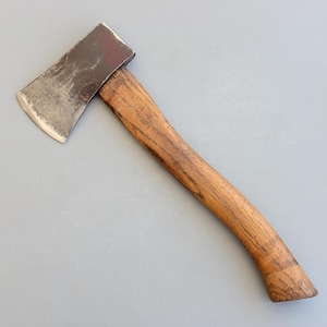 Puede incluir: Un hacha vintage con mango de madera y hoja de acero. La hoja tiene un borde ligeramente curvado y una parte superior pintada de rojo. El mango de madera es de color marrón claro y muestra signos de desgaste.