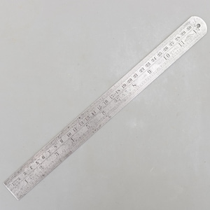 Peut inclure: Une règle en métal argenté avec des marquages en pouces et en centimètres. La règle mesure 30 cm de long. Les chiffres sont gravés dans le métal. La règle est sur une surface grise.