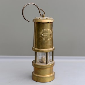 May include: A vintage brass miner's lamp with a curved handle. The lamp has a cylindrical body with a glass section and a circular emblem. The lamp is made of brass and has a weathered appearance.