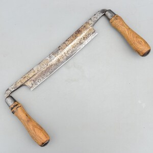 Puede incluir: Una cuchilla de carpintero con una hoja de metal larga y recta que muestra oxidación y desgaste visibles. La herramienta incluye dos mangos de madera unidos a cada extremo de la hoja mediante casquillos metálicos y acabado en madera clara.