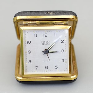 Puede incluir: Un reloj despertador de viaje Europa Deluxe vintage. El reloj tiene una caja dorada con una cubierta de cuero sintético negro. La esfera blanca presenta números y manecillas negras, con las palabras "2 Jewels" y "Germany" visibles.