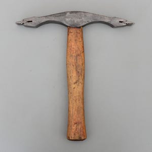 May include: A vintage metal hammer with a wooden handle. The hammer head is a dark grey colour and has two prongs on each end. The handle is a light brown colour and has a textured appearance. The tool is designed for shaping metal.