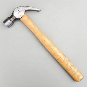 Può includere: Un martello con manico in legno, una tenaglia curva e una testa in metallo. Il martello è etichettato "LEAK PROOF" sulla testa.