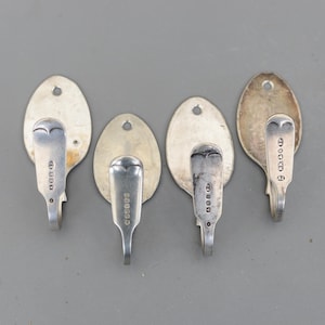 May include: Four silver spoon hooks with oval backplates. Each hook features a spoon bowl design and a curved hook. The backplates have a small hole for mounting. The hooks are arranged on a grey background.