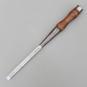 May include: A vintage wood chisel with a metal blade and a wooden handle. The handle is a light brown color and has a metal cap on the end. The blade is silver and has text on it.