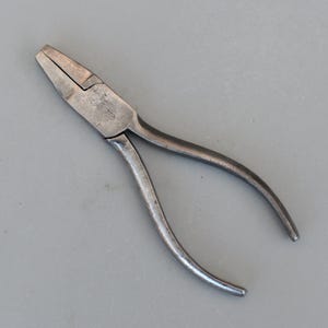 Puede incluir: Un par de alicates de metal plateado, que muestran signos de desgaste. Los alicates tienen una cabeza plana y mangos curvos. La herramienta está diseñada para agarrar, doblar o cortar. El fondo es una superficie gris lisa. Los alicates probablemente se utilizan para diversas tareas.