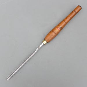 May include: A wood turning chisel featuring a long, polished steel blade and a wooden handle. The handle is light brown with a brass ferrule and a decorative emblem. The tool is designed for shaping wood, commonly used in carpentry.