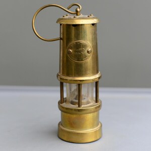 May include: A classic brass miner's lamp, featuring a clear glass section and a curved handle. The lamp has a circular emblem with text. The lamp is cylindrical in shape with a base and top.