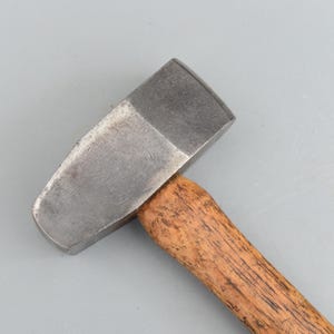 Puede incluir: Un martillo de metal con una cabeza rectangular y un mango de madera. La cabeza del martillo es de color plateado opaco, y el mango es de color marrón claro con vetas de madera visibles. La herramienta está diseñada para golpear y dar forma a materiales.