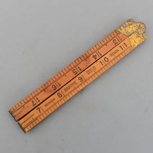 Può includere: Un righello pieghevole in legno vintage con misure in pollici. Il righello è realizzato in bosso e ha cerniere in ottone. Il testo "RABONE" e "MADE IN ENGLAND" sono visibili. Il righello misura circa 30 cm di lunghezza quando è completamente esteso.