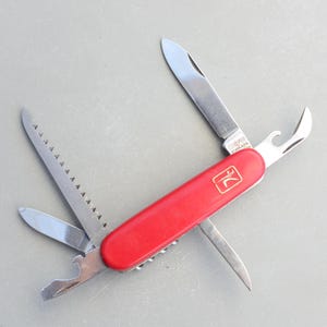 May include: A red multi-tool with various open components, including a knife blade, saw, bottle opener, and small blade. The tool has a red handle with a gold emblem. The stainless steel blades are open against a grey background.