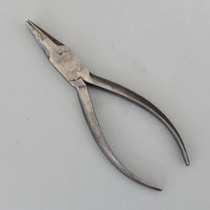 Puede incluir: Un par de alicates de punta larga hechos de metal. Los alicates tienen una punta afilada y mangos curvos. El metal tiene un aspecto desgastado y envejecido. Los mangos tienen un patrón de agarre texturizado.