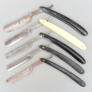 Może przedstawiać: Sześć zabytkowych brzytw z różnymi uchwytami, w tym czarnymi, białymi i z kości słoniowej. Niektóre brzytwy mają wygrawerowane na ostrzu nazwę marki "J.C. Lox & Co. Ground Razor".