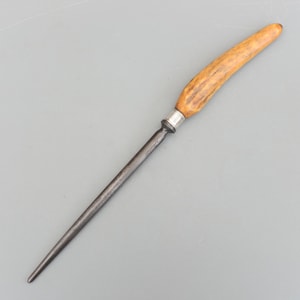 Pode incluir: Um afiador de facas de aço com um cabo texturizado castanho claro. O cabo é conectado à haste de aço com uma peça de metal prateado. O afiador foi projetado para afiar facas e outras ferramentas de corte.