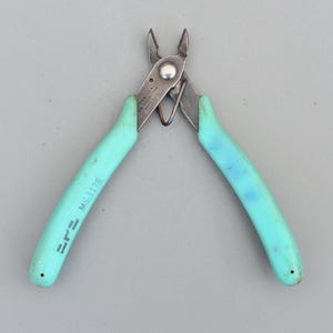 以下が含まれることがあります： ターコイズ色のハンドルと銀色の金属刃が付いたワイヤーストリッパー。ハンドルには「MS 1178」とメーカー名が記載されています。この工具は、電線を切断し、被覆を剥がすように設計されています。