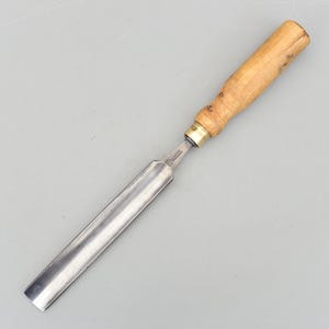 May include: A woodworking chisel with a polished steel blade and a light-coloured wooden handle. The handle is cylindrical and appears to be made of hardwood. The chisel is designed for shaping and carving wood.