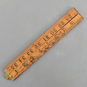 Peut inclure: Une règle pliante en bois vintage avec des marquages noirs. La règle est brun clair avec des signes d'usure visibles et mesure jusqu'à 89 cm. La marque "RABONE" est imprimée sur la règle.