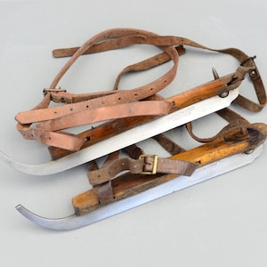 Può includere: Un paio di pattini da ghiaccio in legno vintage con cinturini in pelle. Le lame sono in metallo e le parti in legno sono marroni. I pattini hanno il testo "J.G. SCHROEDER & CO.  CRISTELLER" su di loro.
