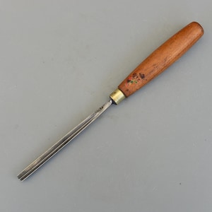 以下が含まれることがあります： 木工用のノミ。木製の柄と金属製の刃が付いています。柄は温かみのある茶色で、真鍮のフェルールと小さな花の模様があります。刃は銀色で、平らで溝のあるデザインです。