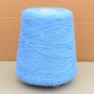 Op de afbeelding: Een grote spoel lichtblauw garen. Het garen is strak gewikkeld rond een conische kern, waardoor een textuuroppervlak ontstaat. De kleur is een zacht, pastelblauw, geschikt voor brei- of haakprojecten. Het garen lijkt pluizig en zacht.