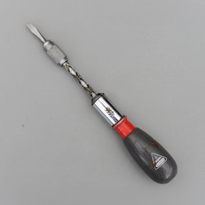 May include: A vintage screwdriver with a flathead tip. The tool features a silver metal shaft with a twisted design, a red band, and a dark gray handle. The handle has a triangular logo and text.