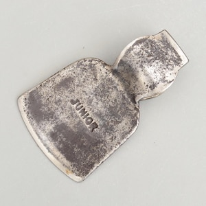 May include: A vintage, weathered metal axe head with the word "JUNIOR" stamped on the blade. The axe head has a curved top and a rectangular blade, showing signs of wear and age.