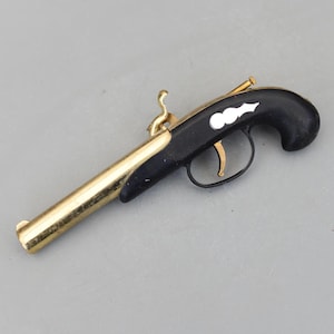 May include: A miniature black and gold pistol with a curved handle and a long barrel. The pistol has a gold trigger, hammer, and barrel, with white decorative accents on the black handle. The design suggests a historical or antique aesthetic.