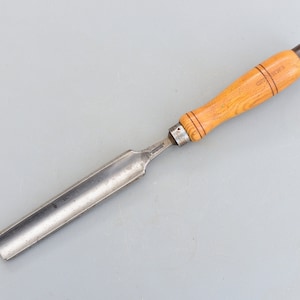 May include: A wooden handled chisel with a metal blade. The handle is made of light wood and has a dark brown stain. The blade is long and thin, and it has a slight curve at the end.