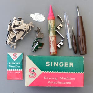 Könnte beinhalten: Eine Sammlung von Vintage-Nähmaschinenaufsätzen und Zubehör von Singer. Enthält eine Schachtel mit Nadeln, eine Schachtel mit Aufsätzen, verschiedene Metallfüße, einen Schraubendreher und eine transparente Ölflasche mit rosa Kappe.