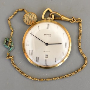 Puede incluir: Un reloj de bolsillo Avia dorado con esfera blanca, manecillas negras y números romanos. El reloj tiene una pantalla de fecha y está sujeto a una cadena y un guardapelo. El reloj dice "AVIA 17 JEWELS INCABLOC SWISS MADE".
