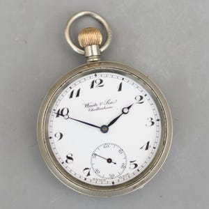 Puede incluir: Un reloj de bolsillo plateado con esfera blanca, números negros y manecillas negras. El reloj tiene un pequeño dial de segundos y el texto "Waite & Son Cheltenham" es visible. La corona de cuerda es dorada.