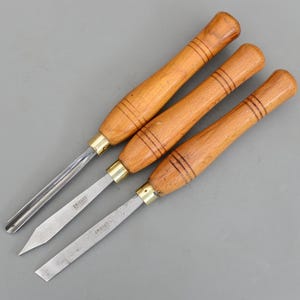 Può includere: Tre scalpelli per tornitura del legno con manici in legno e lame in metallo. I manici sono di colore marrone chiaro con tre anelli marrone scuro. Le lame sono argentate e riportano il marchio "Briggs".