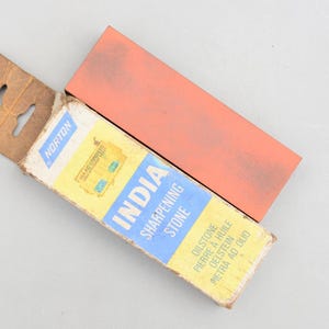 Pode incluir: Uma pedra de amolar retangular laranja ao lado de uma caixa de cartão aberta. A caixa tem um rótulo amarelo e azul com o texto "INDIA SHARPENING STONE" e a marca "NORTON".