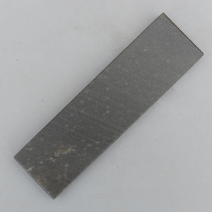 Könnte beinhalten: Ein rechteckiger Schleifstein mit einer dunkelgrauen, feinkörnigen Oberfläche. Der Stein weist subtile, helle Mineralflecken und lineare Streifen auf seiner flachen, matten Textur auf, was auf ein Werkzeug zum Schärfen von Klingen hinweist.