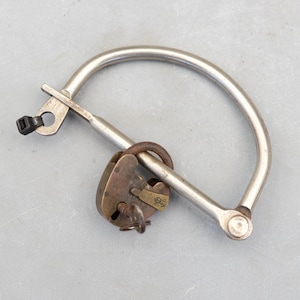 May include: An antique metal shackle with a vintage padlock. The shackle is a curved, silver-colored bar with a connecting mechanism. The padlock is bronze-colored with a key inserted. The items are on a gray surface.