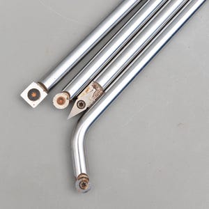 Può includere: Primo piano di diversi utensili per tornio in metallo argentato lucido. Gli utensili hanno varie forme di punta, tra cui quadrata, rotonda e angolata, con dettagli a vite visibili. Gli utensili sono disposti diagonalmente su uno sfondo grigio.