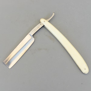 Puede incluir: Una navaja de afeitar abierta, con mango color crema y hoja plateada. La hoja está parcialmente abierta, revelando el filo. La navaja se encuentra sobre un fondo gris.