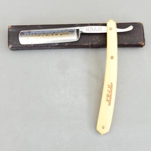 May include: A vintage straight razor with a cream-coloured handle and a silver blade. The blade is marked with the word "KRAM" and the handle also has the word "KRAM" printed on it. The razor is resting on a dark brown case.