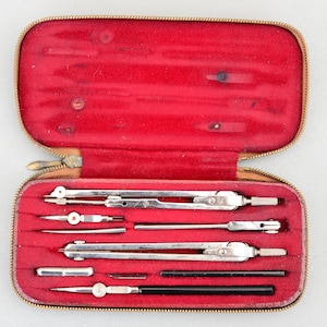 Puede incluir: Un conjunto vintage de herramientas de dibujo en un estuche forrado de terciopelo rojo. Las herramientas incluyen compases, reglas y un lápiz.