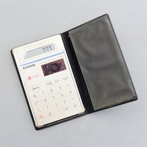 May include: A white Casio SL-900 electronic calculator with a black leather case. The calculator has a small LCD screen displaying "777". The calculator has a variety of buttons, including numbers, operations, and functions.