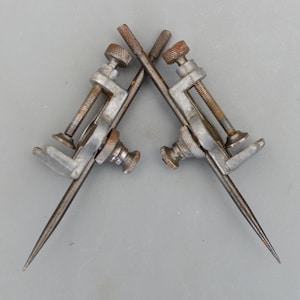 May include: Two vintage metal scribing tools with pointed tips, clamps, and adjustment screws. The tools are arranged in a crossed position, with visible rust on the metal. The background is a plain gray surface.