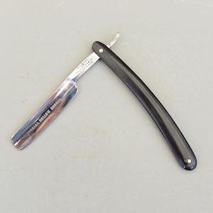 Puede incluir: Una navaja de afeitar recta vintage abierta, con una hoja plateada brillante y un mango negro. La hoja está marcada con las palabras "SPECIAL HOLLOW GROUND". El mango es curvo y tiene un pequeño pasador plateado en cada extremo.