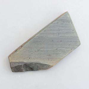 May include: A grey sharpening stone with a rough, textured surface. The stone is rectangular with rounded edges and a slightly irregular shape.